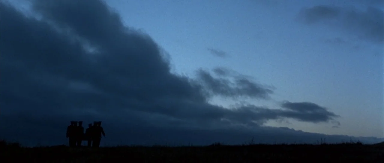 The Rock (1996) movie still: wide — A vast, dark blue sky filled with thick clouds hangs over a grassy field. Four men in uniform walk together…