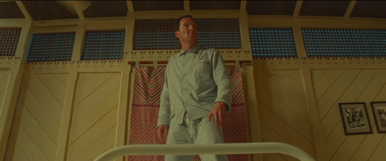 Poison movie still: fullbody — A hospital room with wooden paneled walls and a patterned textile hanging behind a bed. A man in pajamas stands by…