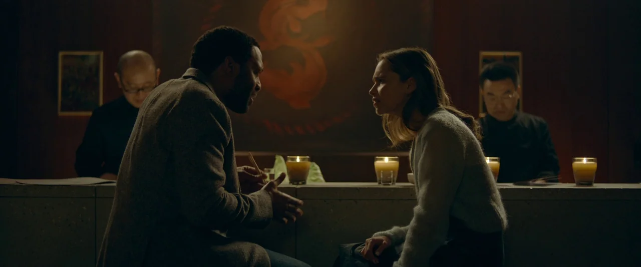 The Pod Generation (2023) movie still: medium — Two individuals converse across a bar counter in a dark, dimly lit environment. Candles placed along…