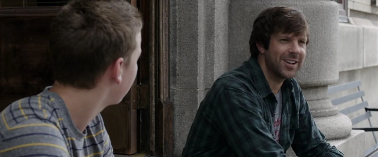 We're the Millers (2013) movie still: medium — Two individuals sit on a concrete ledge outside a building with stone columns. A man engages in…