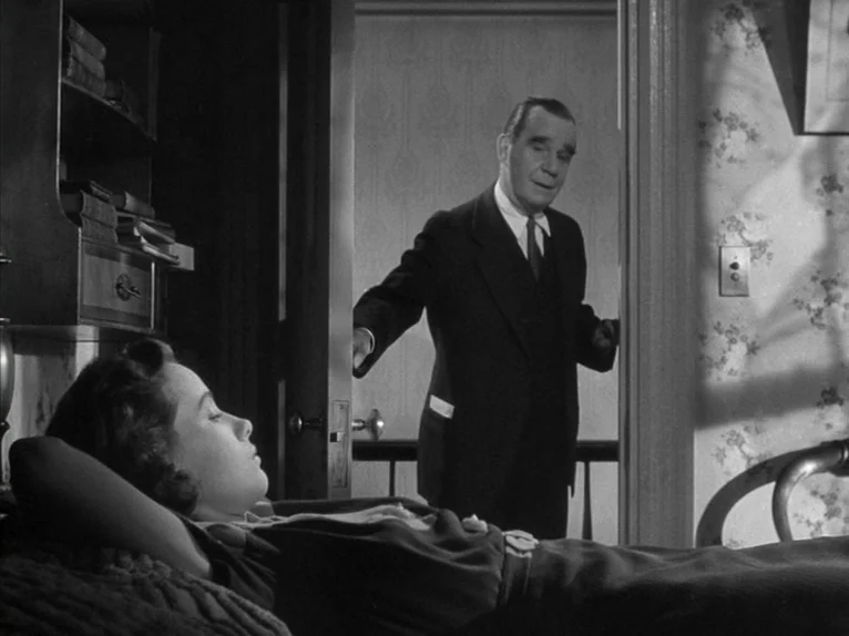 Shadow of a Doubt (1943) movie still: medium — A dimly lit room with floral wallpaper and a small bookshelf integrated into the wall. A woman rests…
