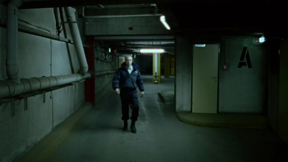 Import Export (2007) movie still: fullbody — A concrete parking garage features exposed piping along walls and an 'A' marked door. A man in a…