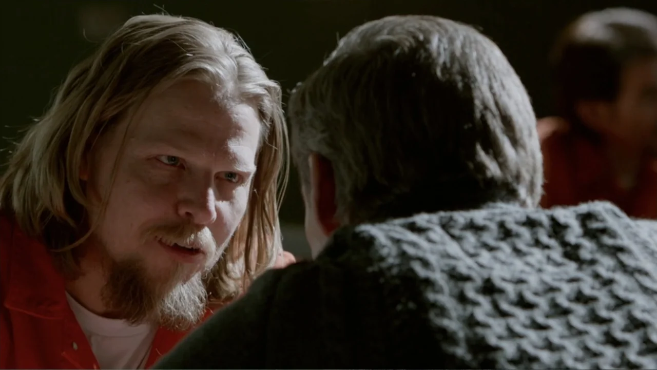 The Captive (2014) movie still: medium — A dimly lit visitation area features two individuals separated by an unseen barrier. A man with long hair…