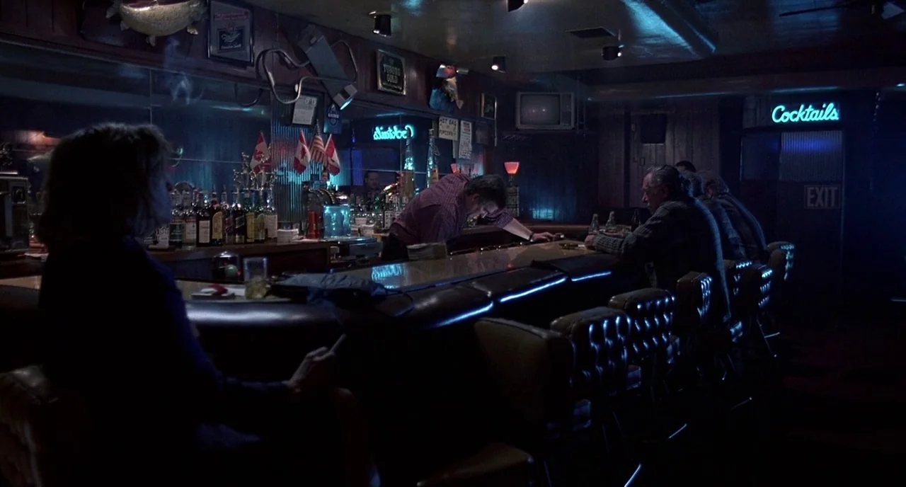 Barfly (1987) movie still: medium — A dimly lit neighborhood tavern with wood-paneled walls and rows of glass bottles. Several patrons sit at the…