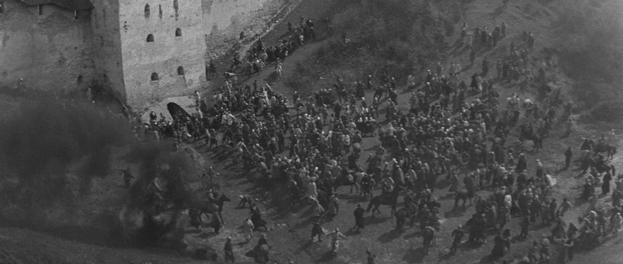 Andrei Rublev (1966) movie still: establishing — An aerial view shows a massive crowd congregating outside the base of a fortress. People and horses…