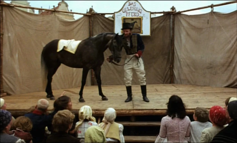 Woyzeck (1979) movie still: fullbody — A wooden platform set up outdoors with canvas backdrops and a painted sign above. A trainer in uniform leads…