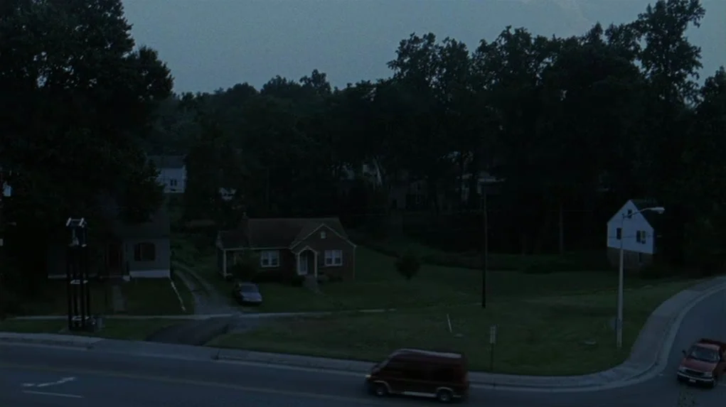 Junebug (2005) movie still: establishing — A suburban residential street at twilight with houses set back from a curved road. A red minivan moves…