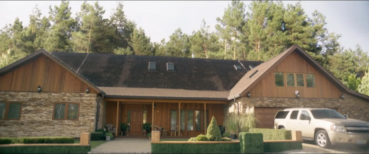 Risqué (2025) movie still: establishing — A sprawling suburban house with wood paneling and stone masonry exterior sits near woods. A tan SUV is…