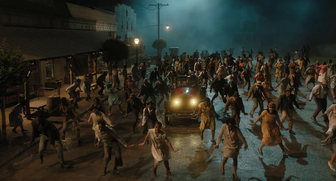 The Color Purple (2023) movie still: wide — A dark, mist-filled main street in a small town features low wooden storefronts. A large crowd of people…