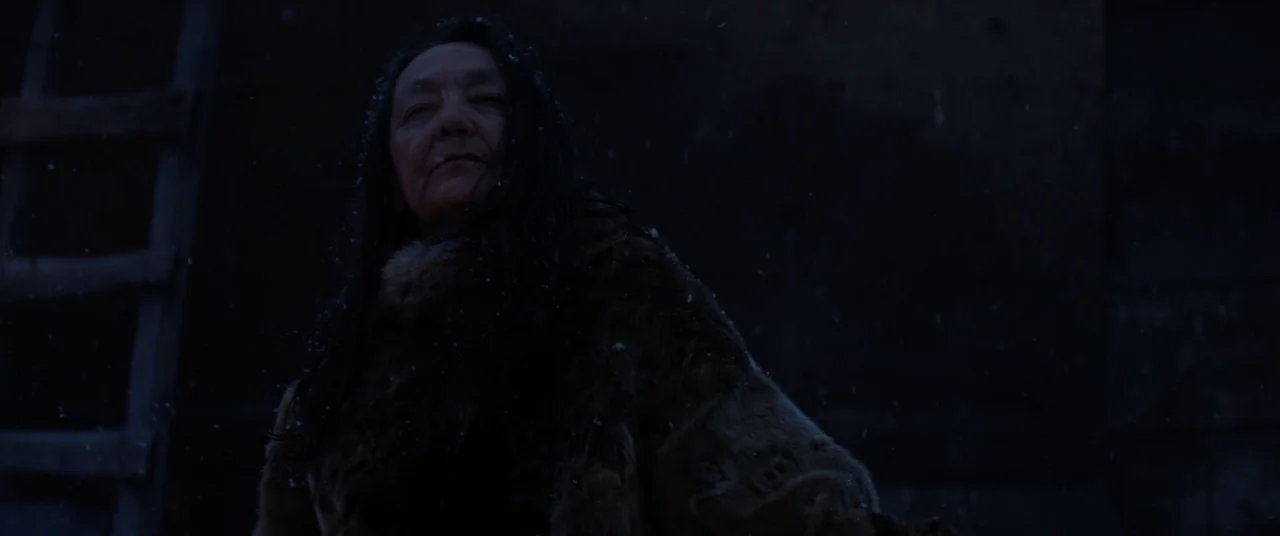 Hold The Dark (2018) movie still: medium — An elderly woman stands outdoors in a dark, snowy environment at night. Light snow falls around her while…