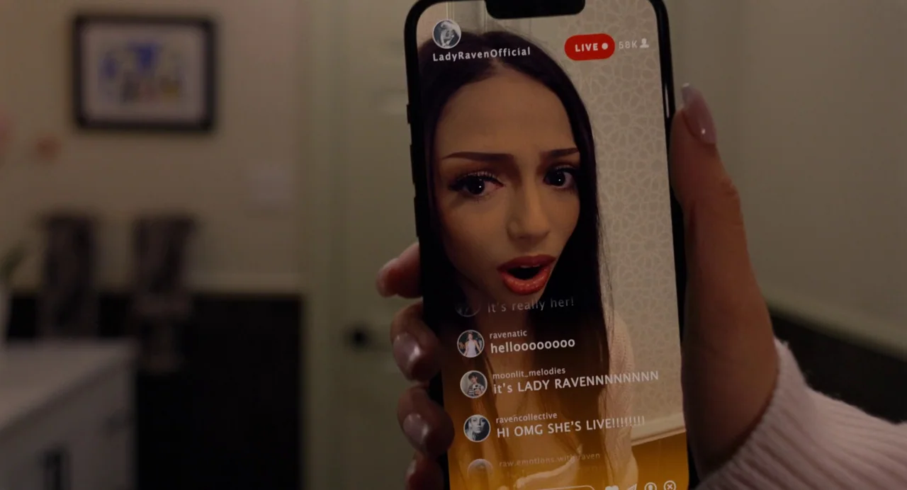 Trap (2024) movie still: closeup — A person holds a smartphone in their hand, displaying a live video stream of a young woman. The background of the…