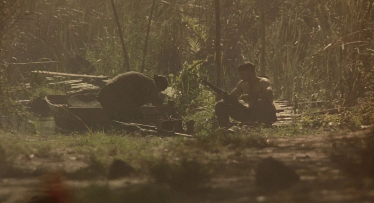 Jacob’s Ladder (1990) movie still: wide — A dense riverbank area features tall reeds and a muddy embankment. Two men prepare equipment next to a…
