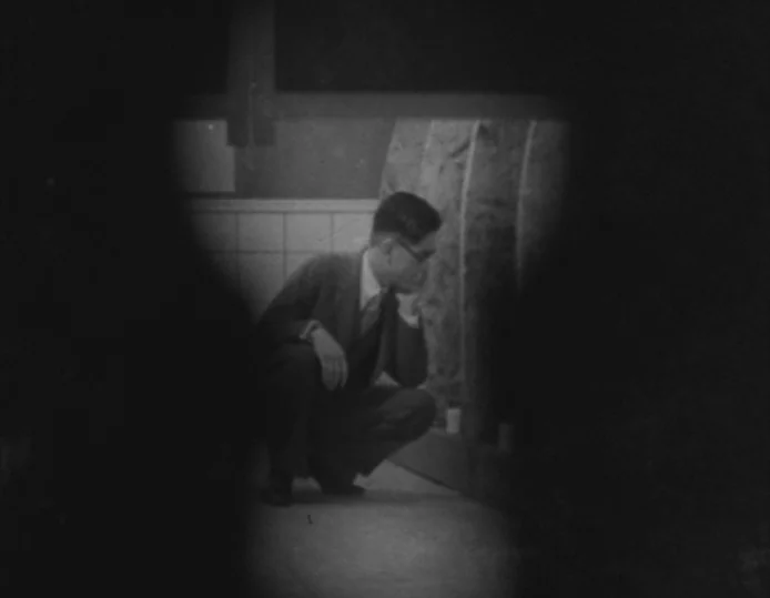 Tokyo Chorus (1931) movie still: medium — A public restroom featuring white tiled walls and a dark concrete floor. A man in a suit crouches near a…