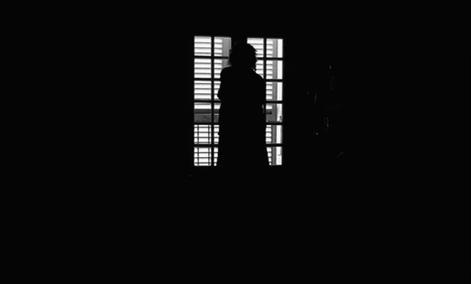 The Man From London (2007) movie still: wide — A dark, minimally furnished room features a singular large glass window with internal shutters. A…