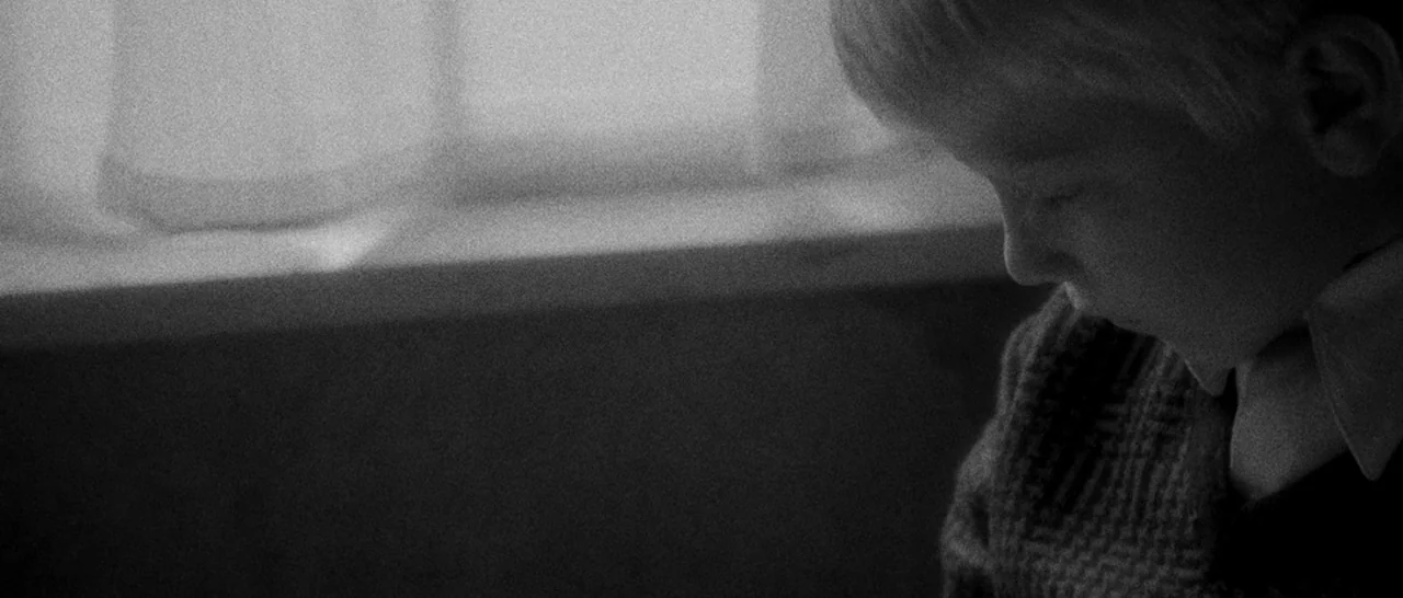 Nymphomaniac Vol 1 (2013) movie still: closeup — A dimly lit interior space features a window with a simple sill and curtain. A young boy with light…