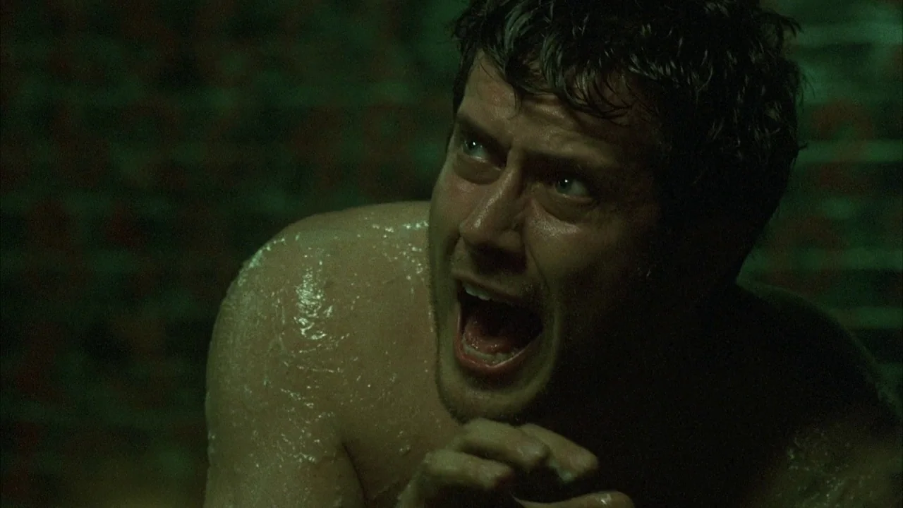 Saw movie still: closeup — A dimly lit, cramped space with dark green-tinted walls and textured surfaces. A shirtless man with sweaty skin yells…