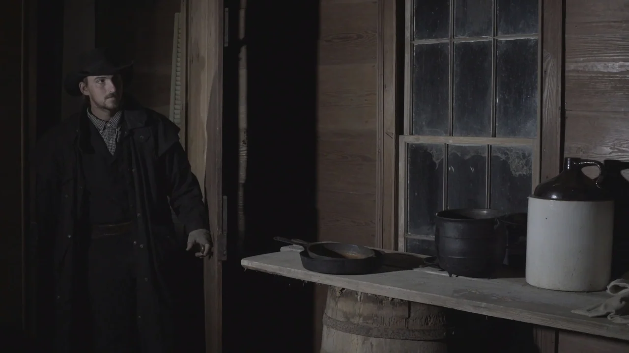 The Dalton Gang (2020) movie still: medium — A rustic wood-paneled room features a workbench with a pot and a ceramic jug. A man in a dark duster…
