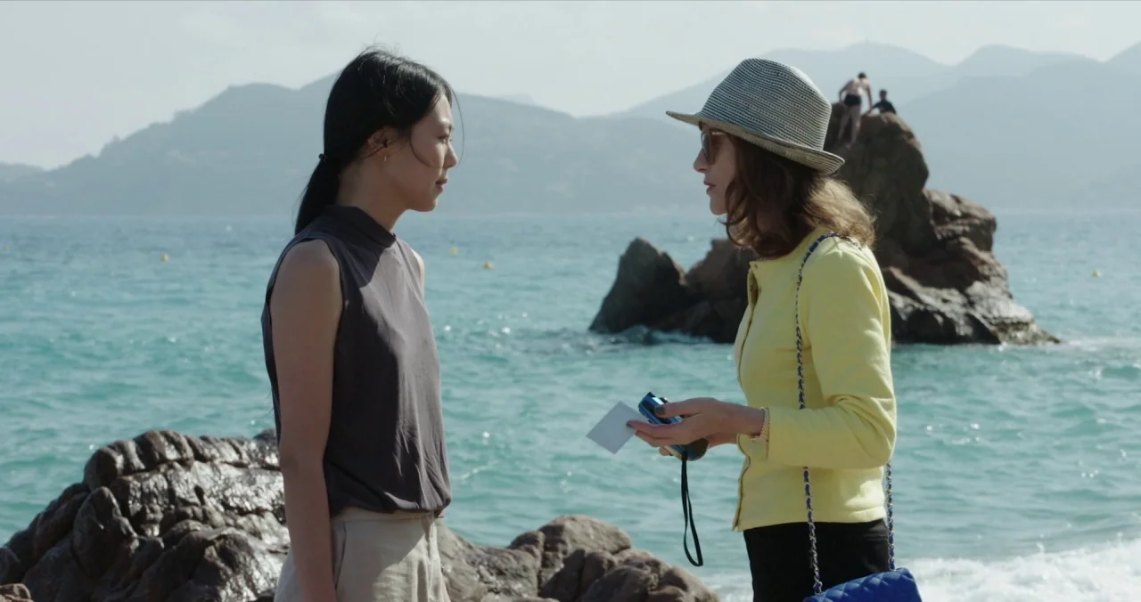 Claire’s Camera (2017) movie still: medium — Two women stand on a rocky shoreline with mountains visible across the ocean background. The woman in…