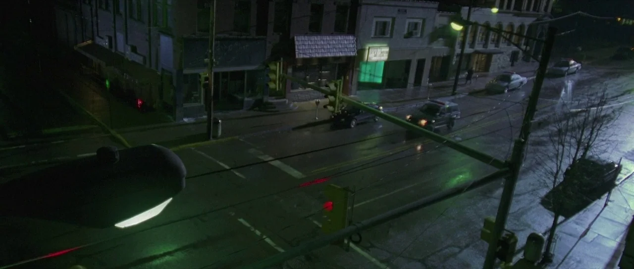The Mothman Prophecies (2002) movie still: establishing — An aerial view of a wet intersection at night with old brick commercial buildings. Several…