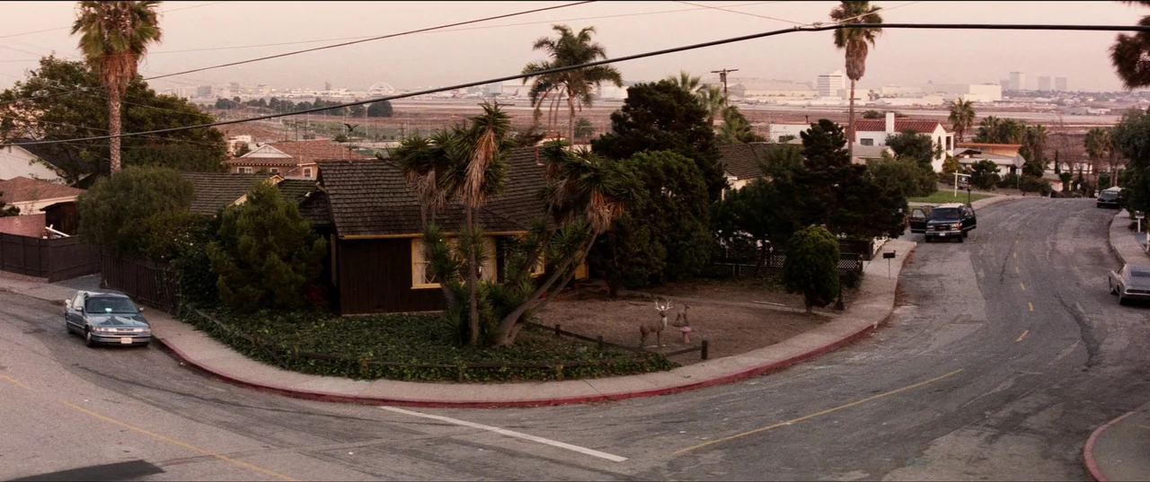 Speed (1994) movie still: establishing — A suburban street corner features a dark wooden house with a patch of dirt. Several parked vehicles sit…