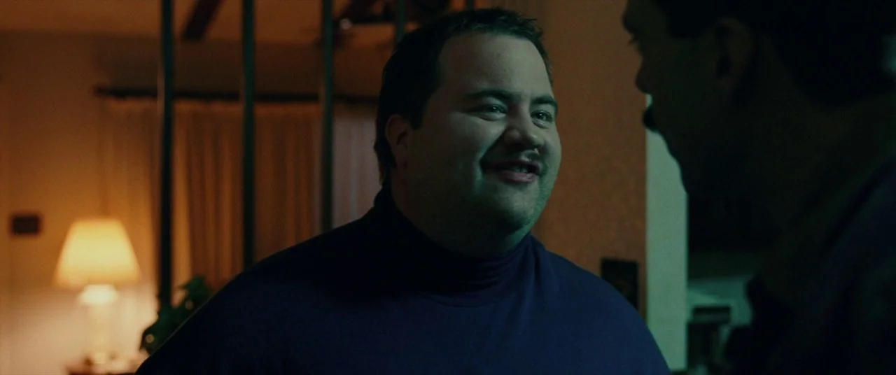 I, Tonya (2017) movie still: medium — An interior space is dimly lit with a table lamp providing the primary light. A man with a mustache faces an…