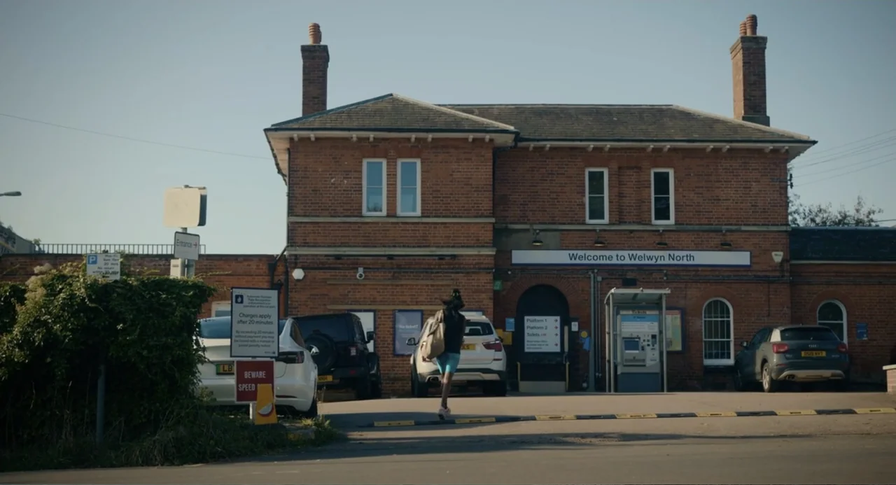 Picture This (2008) movie still: wide — A historic red brick train station building sits at the end of a parking lot. A young woman walks toward the…