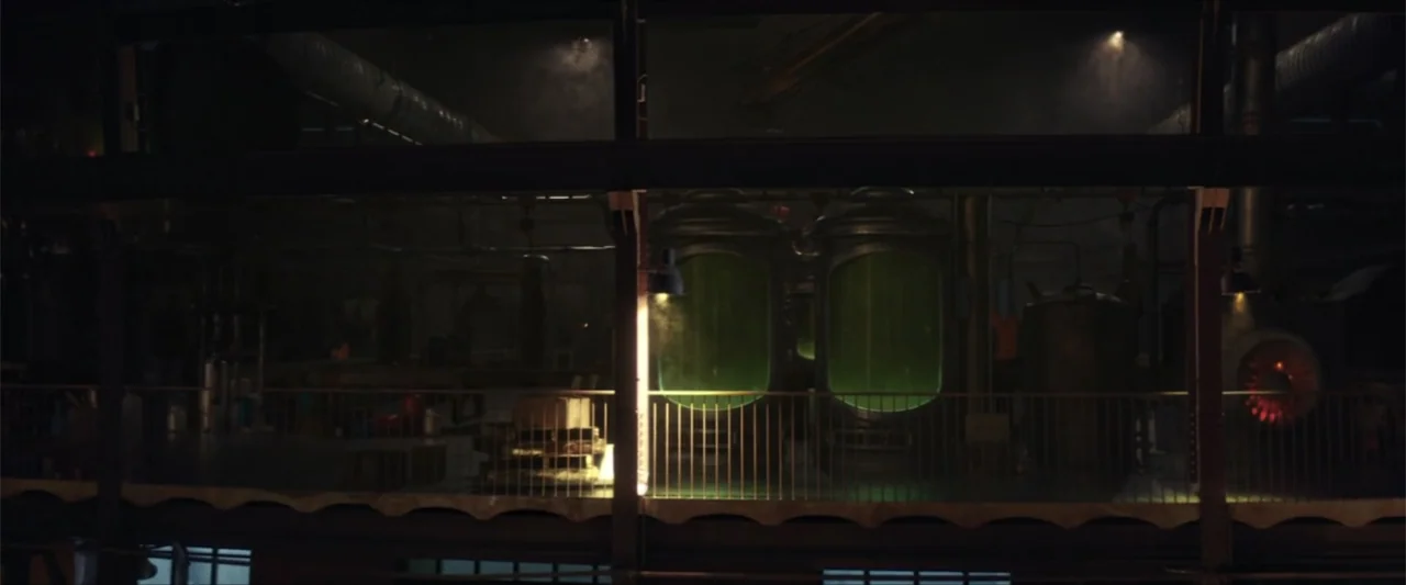 American Carnage (2022) movie still: wide — An industrial laboratory interior features multiple large metal containment vats emitting a green glow.…
