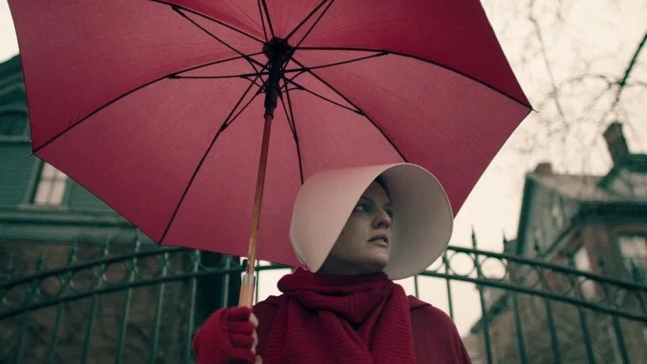The Handmaid's Tale (2017) movie still: medium — A woman stands before a dark wrought iron gate on a cloudy overcast day. She holds a large red…