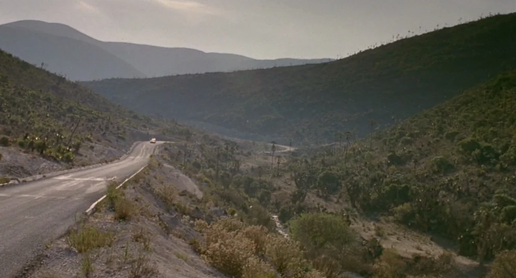 Y Tu Mamá También (2001) movie still: establishing — A long, winding asphalt road cuts through a vast, hilly desert landscape. Sparse vegetation and…