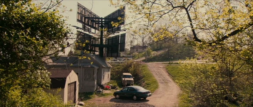 Bubble (2005) movie still: wide — A rural setting features a small house positioned beneath a large elevated metal billboard. A dirt road curves…