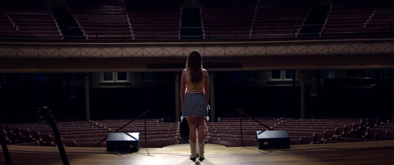 Wild Rose (2018) movie still: fullbody — An empty wooden concert hall features tiered seating under a low balcony in shadow. A woman stands centered…