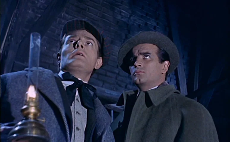 Mill of the Stone Women (1960) movie still: medium — A cramped, dark wooden attic space is visible behind two men standing in darkness. They hold a…
