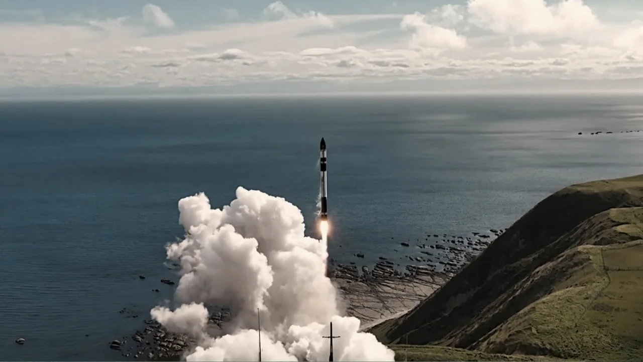 Wild Wild Space (2024) movie still: wide — A tall thin rocket lifts off from a rocky coastline into the sky. Dense white exhaust smoke fills the…