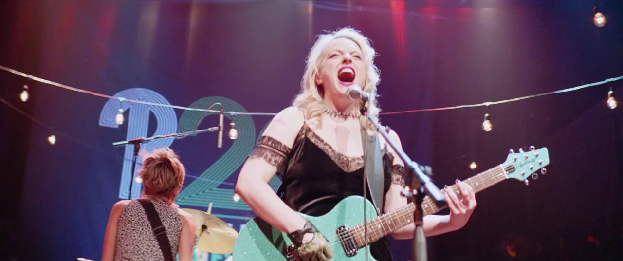 Her Smell (2018) movie still: medium — A dark concert hall features a band performing under strings of hanging light bulbs. The lead singer belts…