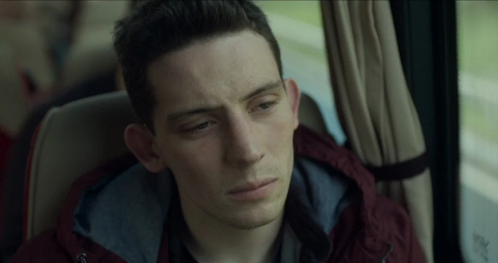 God’s Own Country (2017) movie still: closeup — The interior of a passenger bus with beige headrests and window curtains. A young man sits in a…