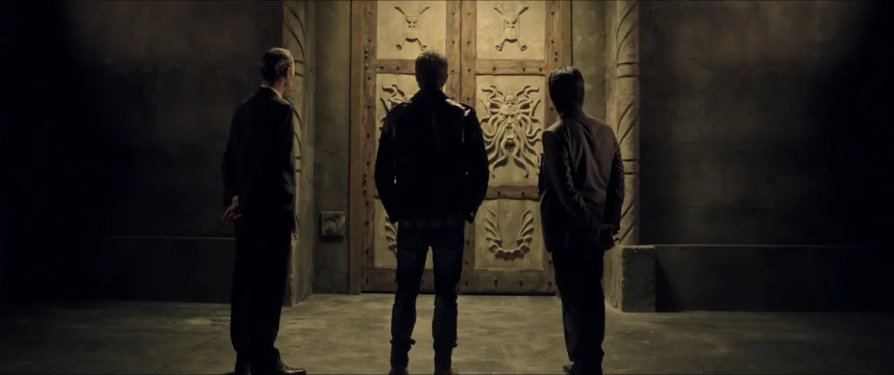 Room 6 (2006) movie still: fullbody — A dimly lit stone room features a massive wooden door carved with an intricate relief. Three men stand in a…