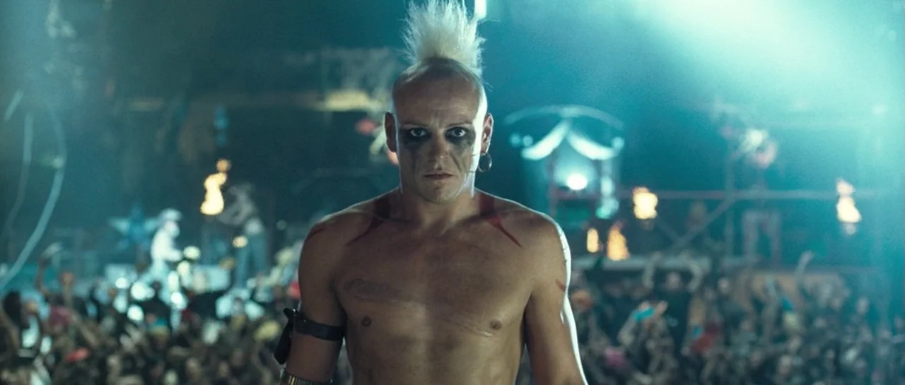 Doomsday (2008) movie still: medium — An open air concert venue with a large crowd gathered under dramatic stage lights. A shirtless man with a…
