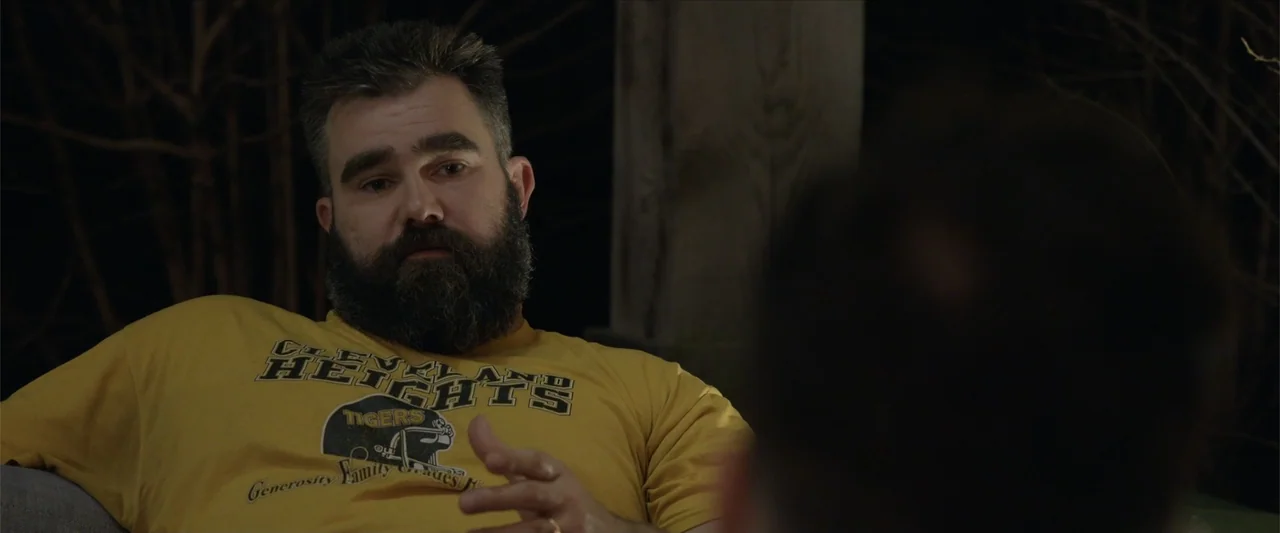 Kelce (2023) movie still: medium — A dark outdoor setting at night with a person seated in the foreground. A man with a dark beard wearing a yellow…
