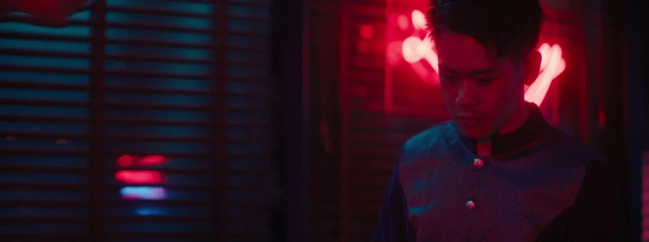 Dead Pigs (2018) movie still: medium — A dark space features a bright red neon light reflecting off a nearby surface. A man in a patterned shirt…