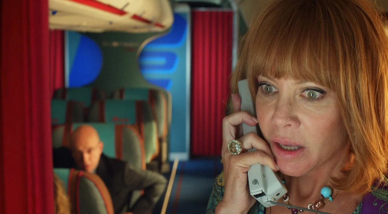 I’m So Excited! (2013) movie still: closeup — A retro-styled airplane interior features red curtains and teal passenger seats. A woman in the…