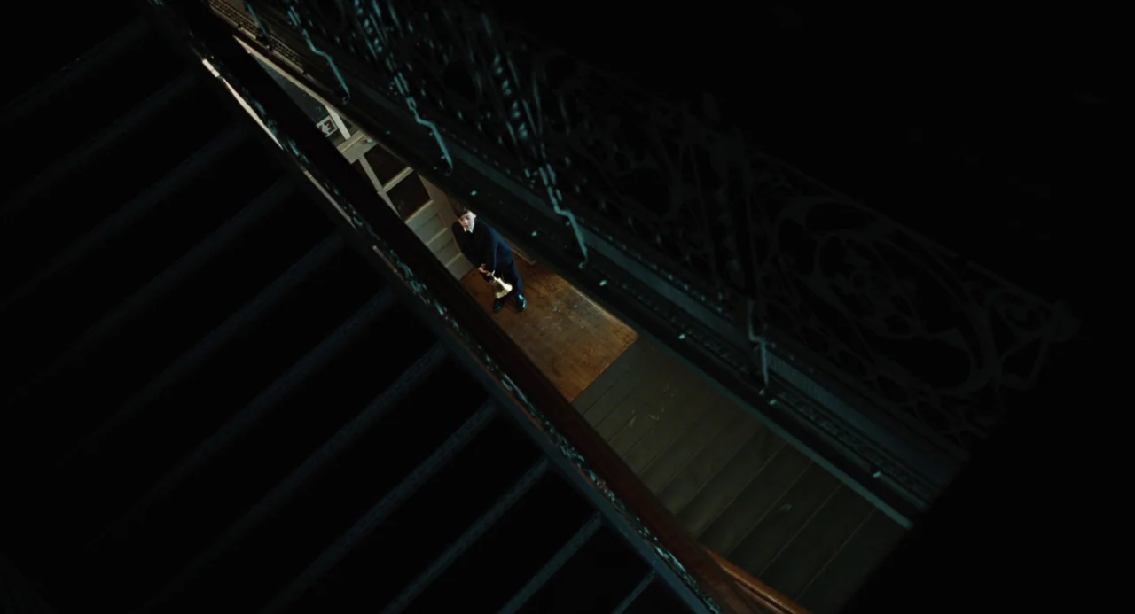 Doubt (2008) movie still: wide — A dark, grand staircase interior with ornate metal railings and dim wooden steps. A young boy in a dark suit stands…