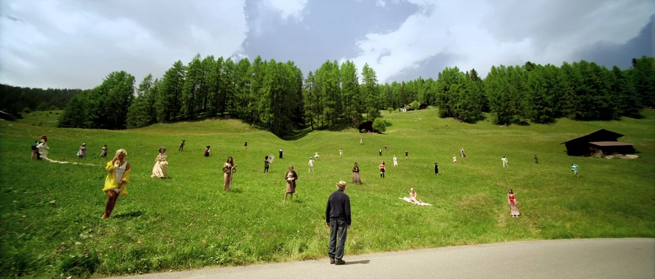 Youth (2015) movie still: wide — A vast green mountain meadow features a line of tall pine trees and a hut. Numerous people in varied outfits stand…