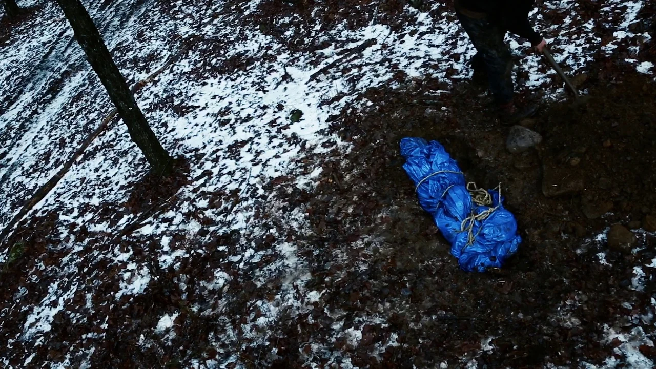 The Deeper You Dig (2019) movie still: wide — A patch of muddy ground covered in fallen leaves and light snow in woods. A person digs a hole in the…