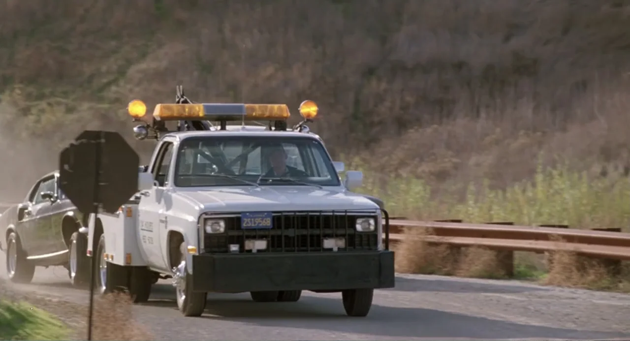 Dangerously Close (1986) movie still: medium — A white tow truck travels along a road with dry grass and metal guardrails. A person sits in the…
