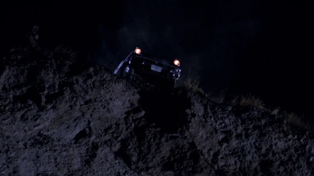 Cronos (1993) movie still: wide — A dark, uneven dirt hillside rises sharply against a black night sky. A vintage car rests near the top ridge with…