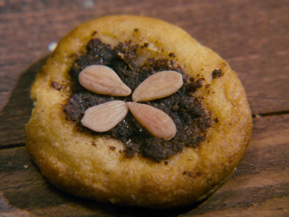 Alice (1988) movie still: closeup — A small round cookie with a dark filling sits on a dark wooden surface. Four blanched almond slivers are…