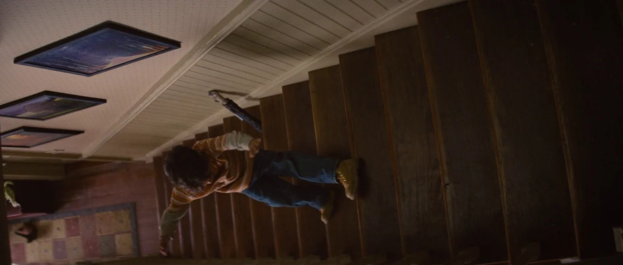 Looper (2012) movie still: fullbody — A residential staircase featuring dark wooden steps and light wallpaper with framed wall art. A young boy in…