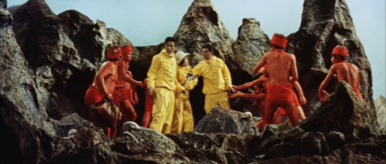 Mothra Vs Godzilla (1964) movie still: wide — A group of people wearing bright yellow jackets stands amidst jagged, desolate stone outcroppings.…