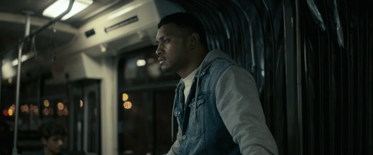 Talk to Me (2023) movie still: medium — A dimly lit interior of a public bus at night with dark exterior windows. A man stands and looks out the…