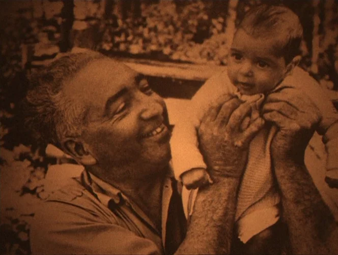 WR: Mysteries of the Organism (1971) movie still: medium — A man holds a small baby up in the air in an outdoor setting. The background is composed…
